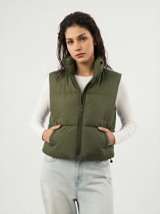 Woman wearing an olive green puffer vest over a white long-sleeve shirt on a light gray background