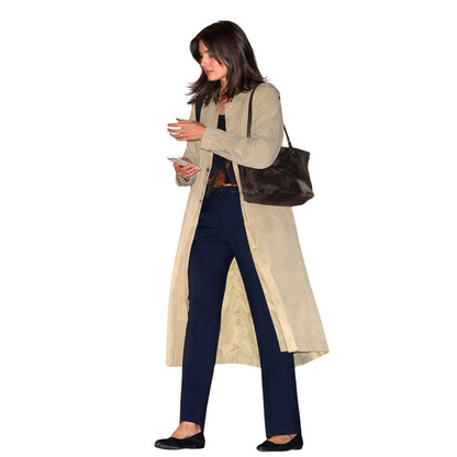 Woman in a beige trench coat and navy pants holding a phone and a brown handbag on a white background