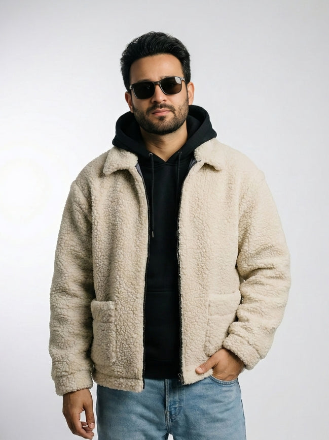 Man wearing a beige sherpa jacket, black hoodie, sunglasses, and blue jeans on a light gray background