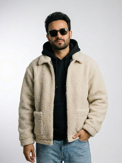 Man wearing a beige sherpa jacket, black hoodie, sunglasses, and blue jeans on a light gray background