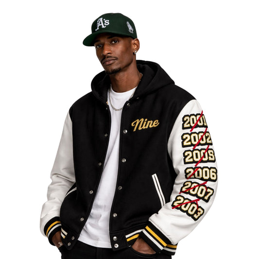 Man wearing a black and white letterman jacket with 'Nine' and years printed on sleeves, paired with a green baseball cap.