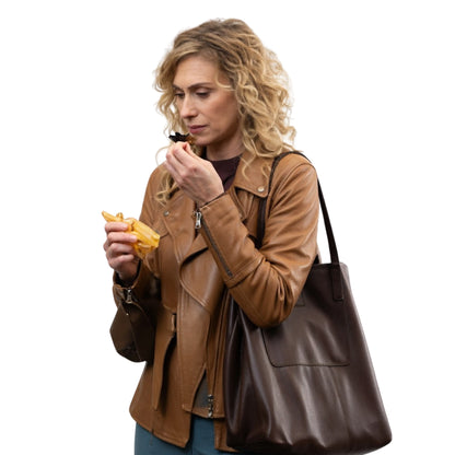 Woman in a brown leather jacket holding a brown leather handbag and eating a snack.